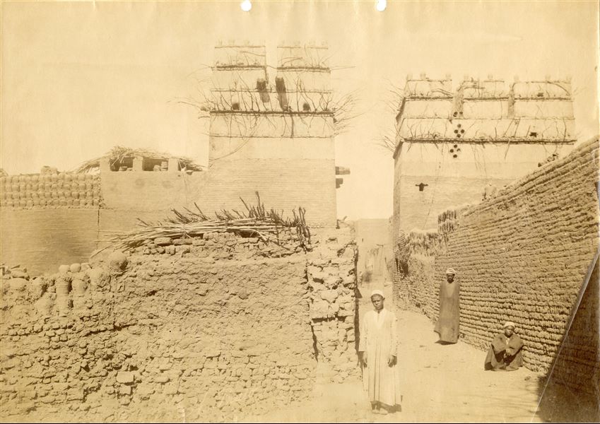The photograph shows a view of a street in an Egyptian village (difficult to identify) built of mud brick. Clay pots can be seen in the top left corner. A man and two young boys, looking at the camera lens have also been photographed. The author's signature albeit faded and almost illegible, is at the bottom. 