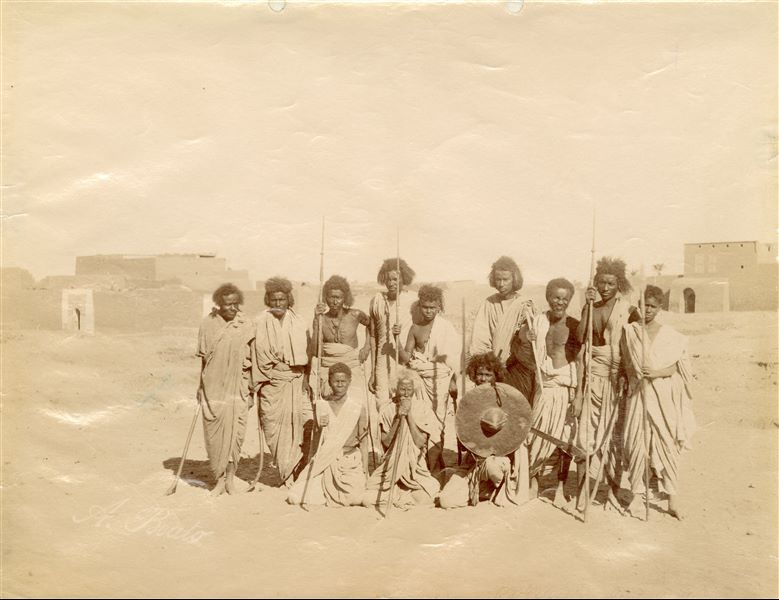 Popolazione nubiana in posa per uno scatto fotografico, presumibilmente dei guerrieri o cacciatori: alcuni tengono in mano lance e spade, un giovane mostra invece uno scudo. In basso a sinistra la firma poco visibile dell’autore.