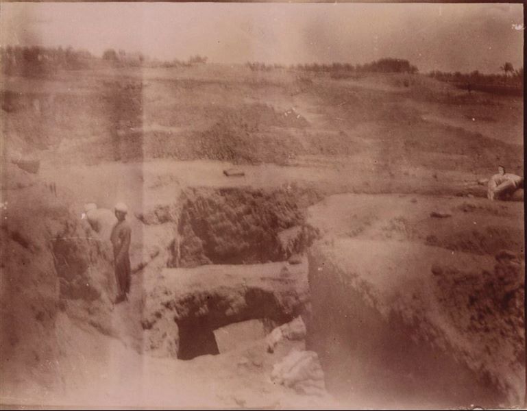 Excavation trench in the area of the Kom, where a bridge structure can be seen. In the background, the cultivated fields of Heliopolis can be seen, as well as a house and a series of trees. Photograph quite blurred and unfortunately not very sharp. Schiaparelli excavations.