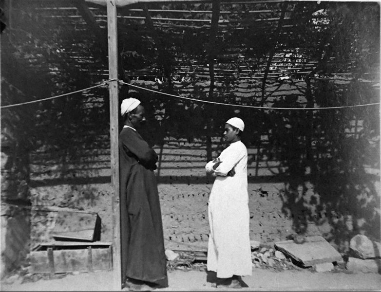 Photograph of two members of the mission, on the left the cook Atallah and on the right is the young Buhus, both standing under a veranda of the house in Heliopolis. Angelo Sesana Archive. 