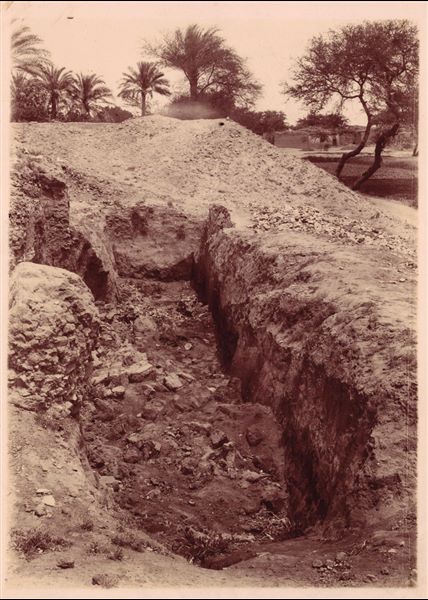 Photograph of an excavation trench, with some blocks surfacing from the earth. In the background, there are some dwellings. Schiaparelli excavations. 