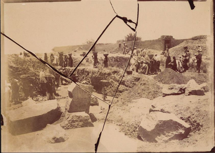 Temple of Mnevis, trench with fragments of the naos of Psamtik I, close to housing. Note the muddy ground. This paper print has been toned towards sepia. The plate (from which the print was teken) was broken (as can be seen from the print itself) and unfortunately no longer exists. Schiaparelli excavations. 