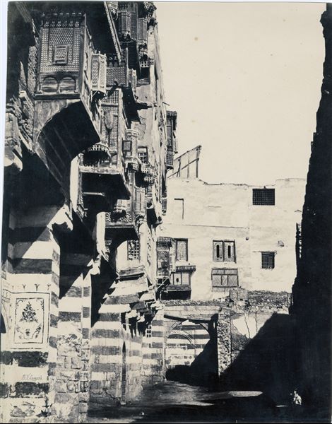 View of a street in Cairo, showing houses with alternating light coloured and red bricks, also featuring mashrabiyas, the characteristic balconies with ornate wooden grates. The author's signature can be found at lower left. 