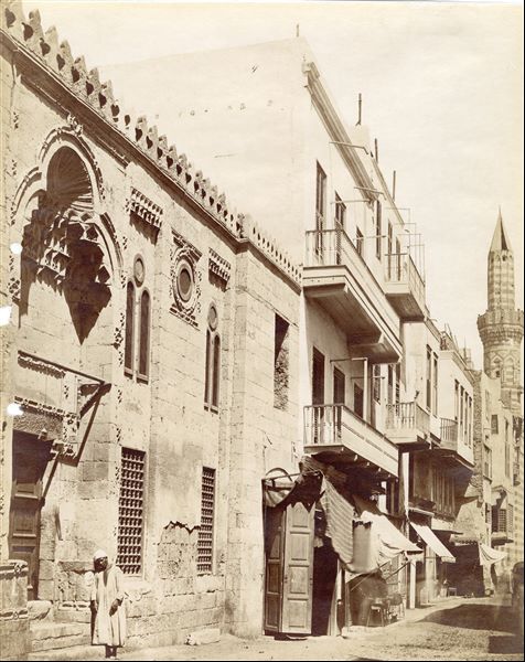 This shot provides a glimpse of a street (presumably) in Cairo, with an Egyptian man posing next to the elaborate entrance of a building. Along the street, the minaret of a mosque.  