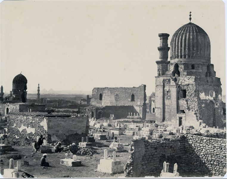 Islamic cemetery in Cairo, where some structures including domes can be seen in a state of disrepair and in danger of collapsing. In the background, the silhouettes of two of the three Great Pyramids of Giza can be seen. The author’s signature is slightly visible at the bottom centre. 
