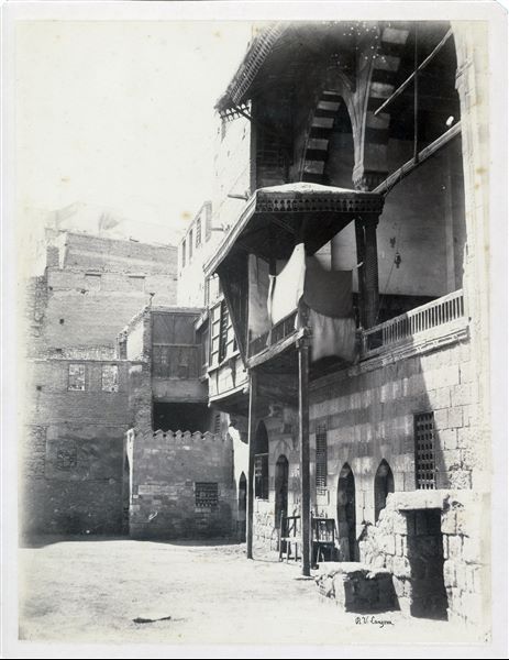Detail of a street in the city of Cairo, where mashrabiyas (characteristic balconies) are visible. On the right there is an arched arcade with alternating light coloured and red stones, typical of Islamic art. The author's signature is at bottom right.