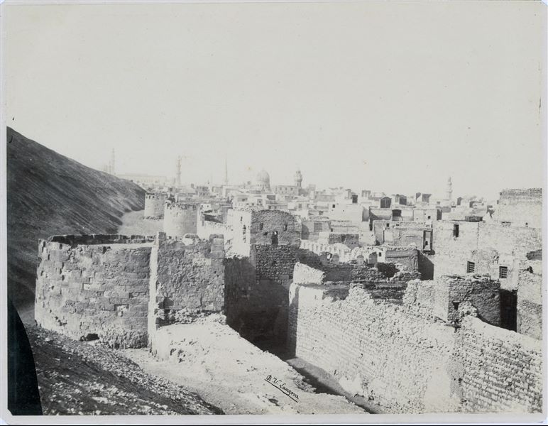 Panorama della città del Cairo, nel quale si intravedono, sullo sfondo, alcune moschee e minareti. In primo piano, resti delle mura medievali, come da indicazioni nel retro della stampa. La firma dell’autore si trova in basso al centro. 