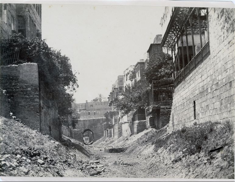 Glimpse of a street in Cairo, leading to a small gate in the city walls. Some wooden pergolas are visible near the dwellings. Note that the level of the street is lower than that of the houses, to reach them one had to use narrow steps. The author's signature can be seen at the bottom centre. 