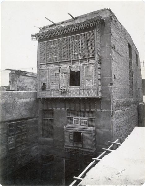 Fotografia di un’abitazione egiziana presumibilmente al Cairo. Essa mostra su tre piani le mashrabiyye, i caratteristici balconi chiusi con grate di legno decorate. La firma dell’autore si trova in basso a destra.