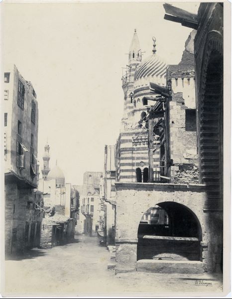 View of a street in Cairo. In the background on the left, the minarets of the Al-Rifa'i Mosque, which houses the mausoleum of part of Mohammed Ali's family and successors, including 20th century rulers including King Fuad I and King Farouk, can be seen. The author's signature is visible at the bottom right-hand corner. 