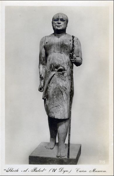 Gallery in the Egyptian Museum in Cairo. Photograph of the famous wooden statue of the official Kaaper (4th-5th dynasty, Cairo CG 34), commonly known as Sheik el-Beled. During excavations of his tomb at Saqqara, conducted by Mariette, the workers who unearthed this statue christened it with the name Sheikh-el-Beled, meaning “village chief” in Arabic due to its resemblance to a local village chief at the time. This name has remained in use until today. Album “Cartes postales” (Postcards).