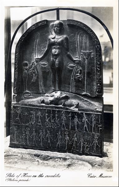 Gallery in the Egyptian Museum in Cairo. A stele from the Ptolemaic Period depicting a young god Horus standing on top of two crocodiles, holding four snakes and other animals: a lion, a goat and two scorpions. Album “Cartes postales” (Postcards).