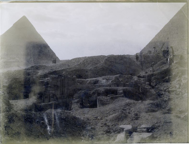 Fotografia di scavo della tomba a mastaba del dignitario Iteti. La fotografia mostra in secondo piano le due grandi piramidi, quella di Cheope a destra, e di Chefren a sinistra. L’immagine purtroppo risulta leggermente chiara ai margini, impedendo una completa comprensione del documento. Scavi Schiaparelli. 