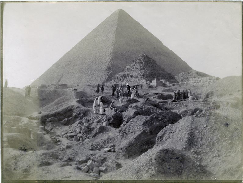 Fotografia di scavo dell’area dove è situata la tomba a mastaba del dignitario Iteti. La fotografia mostra in secondo piano la piramide di Cheope, insieme a due delle piramidi satellite. In questo scatto lo scavo non ha ancora portato alla luce la facciata della mastaba, si vedono diversi operai al lavoro e probabilmente, al centro, Evaristo Breccia mentre dirige i lavori. L’immagine risulta leggermente chiara ai margini. Scavi Schiaparelli. 