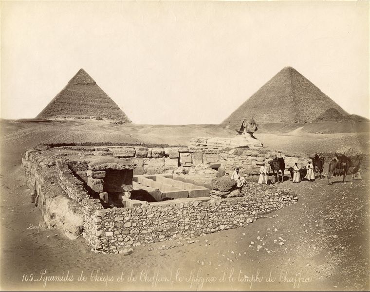 The photograph shows, in the foreground, some Egyptians resting with four camels at the Valley Temple of the pyramid complex of Khafre. In the background the sphinx is visible and already partially cleared of sand, it is thought to be commissioned by Pharaoh Khafre. In the background are the pyramids of Khafre (left) and Khufu (right), pharaohs of the 4th dynasty. The author's signature can be found at the bottom left.  