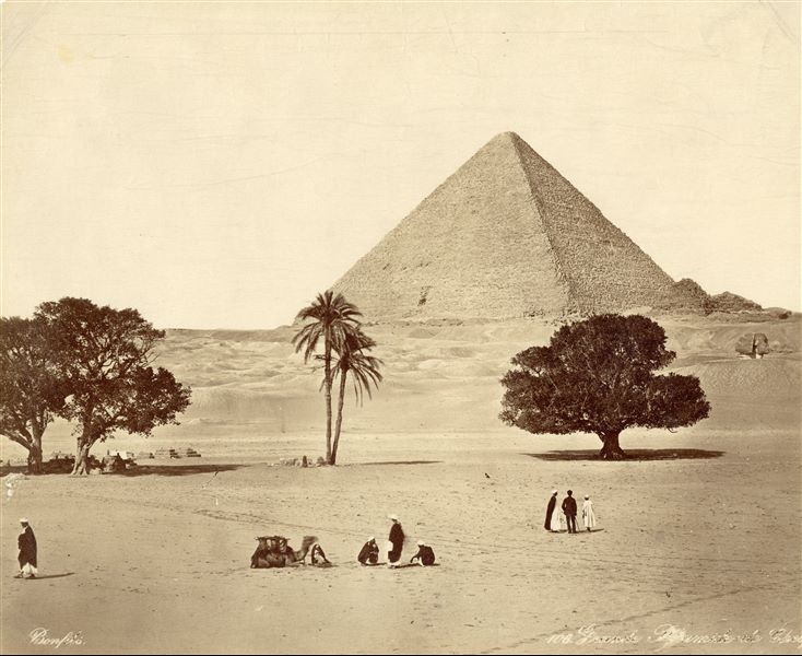 The pyramid of Pharaoh Khufu and its satellite pyramids form the photograph’s backdrop, which depicts a part of the Giza Plateau where some trees are also visible. Some people, including one in Western clothing with a staff, are observing the view, where the head of the Sphinx (on the right) also stands out. Some of these figures, as well as the camel are slightly out of focus. The photographer's signature is visible at the bottom left.