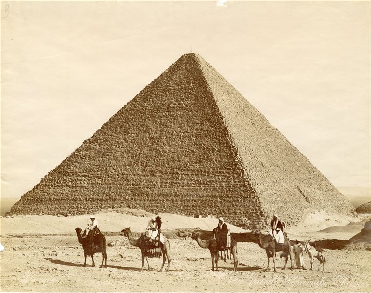 Veduta della piramide di Cheope, la più grande delle tre piramidi di Giza, davanti alla quale sono in posa alcune guide locali. Sulla destra è appena visibile una piramide satellite. Lo scatto è firmato in basso a sinistra.