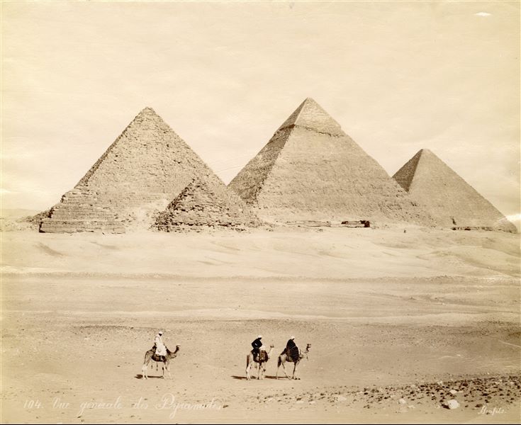 Veduta delle piramidi di Giza con tre beduini in groppa a dromedari in posa per lo scatto. A sinistra si riconosce la piramide di Micerino, la più piccola delle tre, con due piramidi satellite. Al centro vi è la piramide di Chefren e a destra quella di Cheope. La firma del fotografo è posta in basso a destra.
