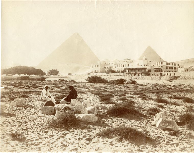 The photograph depicts two Egyptians in the foreground sitting on some large boulders, possibly fragments from antiquity, in front of the Mena House Hotel, which had already been extended with its upper floors and dates the photograph to after 1883. In the background the pyramids of Khufu (centre) and Khafre (right) are visible. The author's signature can be seen at the bottom left. 