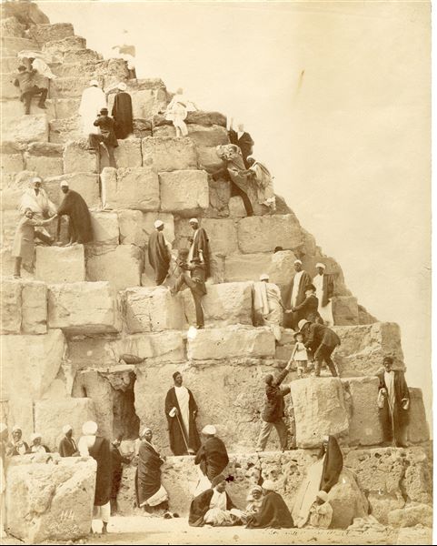 The photograph shows tourists being helped by locals, attempting to climb the pyramid of Khufu at Giza. The author's signature is at the bottom left.