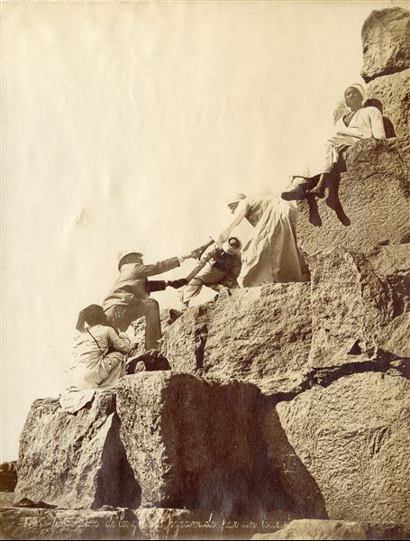 Three egyptian guides help a tourist climb the pyramid of Pharaoh Khufu, while a fourth egyptian is seated on the blocks higher up. There is no signature of the author, although the work could be attributed to F. Bonfils.