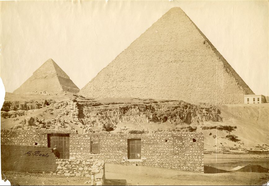 Le piramidi dei faraoni Chefren e Cheope sovrastano la piana di Giza, dove si trovano alcune costruzioni moderne. In basso a sinistra la firma dell’autore.