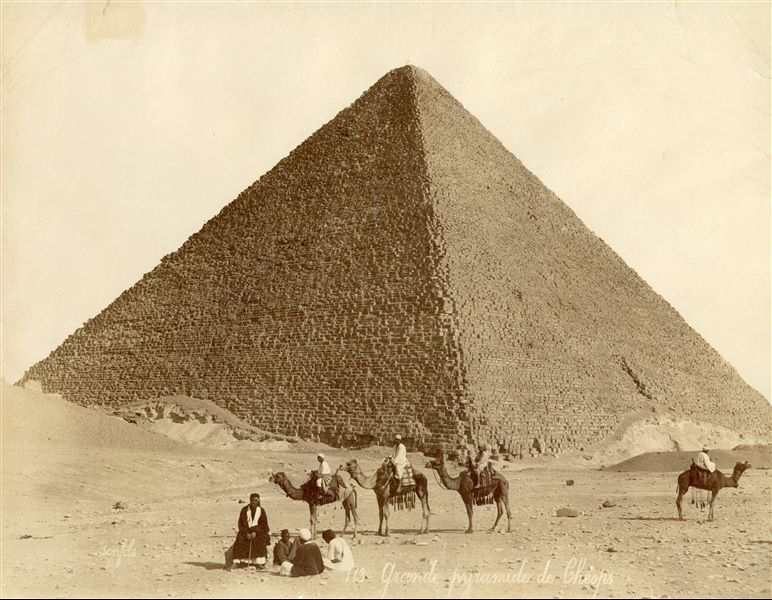 Fotografia della piramide di Cheope, faraone della IV Dinastia, a Giza. Ai piedi della piramide, sono presenti otto abitanti locali, alcuni seduti, altri su dromedari. In basso a sinistra vi è la firma dell’autore.