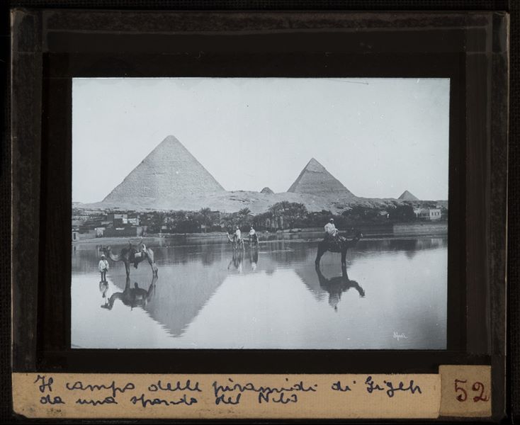 Piramidi di Giza viste dall'altra sponda di un ramo del fiume, dove si trovano in posa alcuni egiziani con i propri cammelli. In basso a destra si riconosce la firma dell'autore, Bonfils, scritta al contrario.