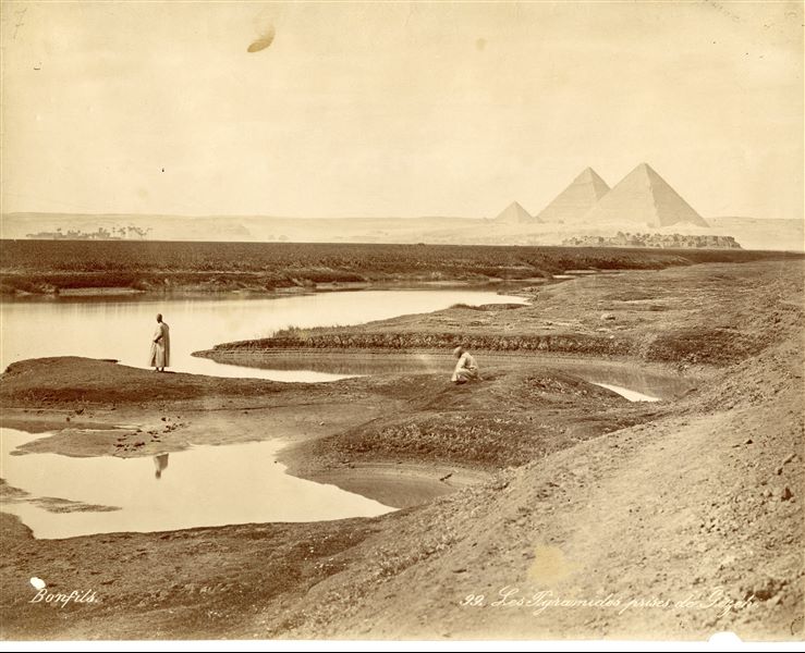 Photograph of the Giza Plateau, showing, in the foreground, the retreating body of water after a flood and two Egyptians monitoring the situation. In the background, the three great pyramids, of Khufu, Khafre and Menkaure, dominate the plain. The author's signature is present at the bottom left.