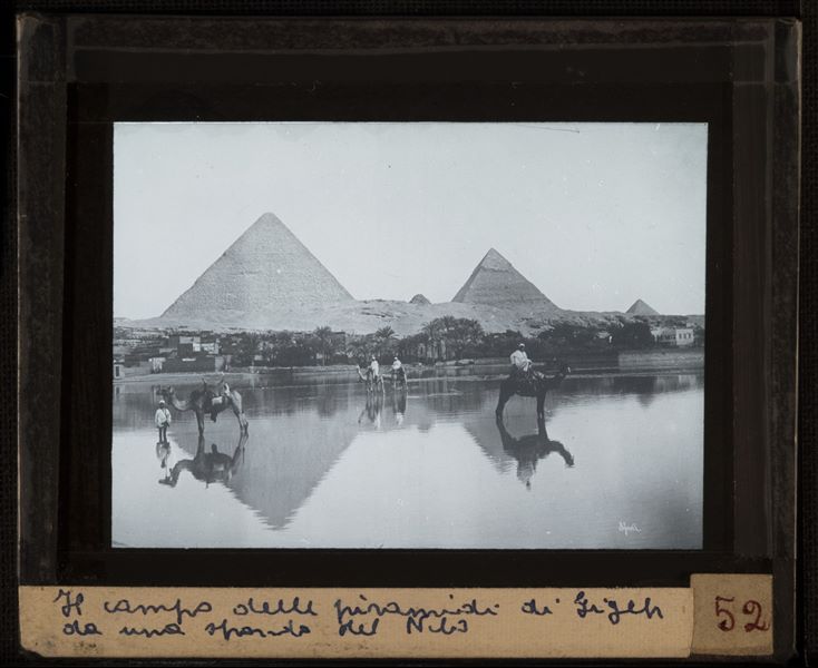 Pyramids of Giza seen from the other side of the riverbank, where some Egyptians are posing with their camels. In the bottom right-hand corner, the signature of the author Bonfils is visible, written in reverse.  