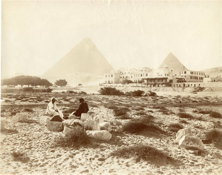 The photograph depicts two Egyptians in the foreground sitting on some large boulders, possibly fragments from antiquity, in front of the Mena House Hotel, which had already been extended with its upper floors and dates the photograph to after 1883. In the background the pyramids of Khufu (centre) and Khafre (right) are visible. The author's signature can be seen at the bottom left. 