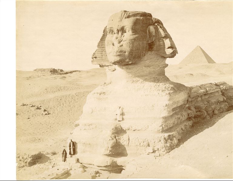The photograph shows the Sphinx of Giza, with an Egyptian climbing the monument and two other people at its base. In the background, the pyramid of Menkaure can be seen. The author's signature is visible at the bottom left.