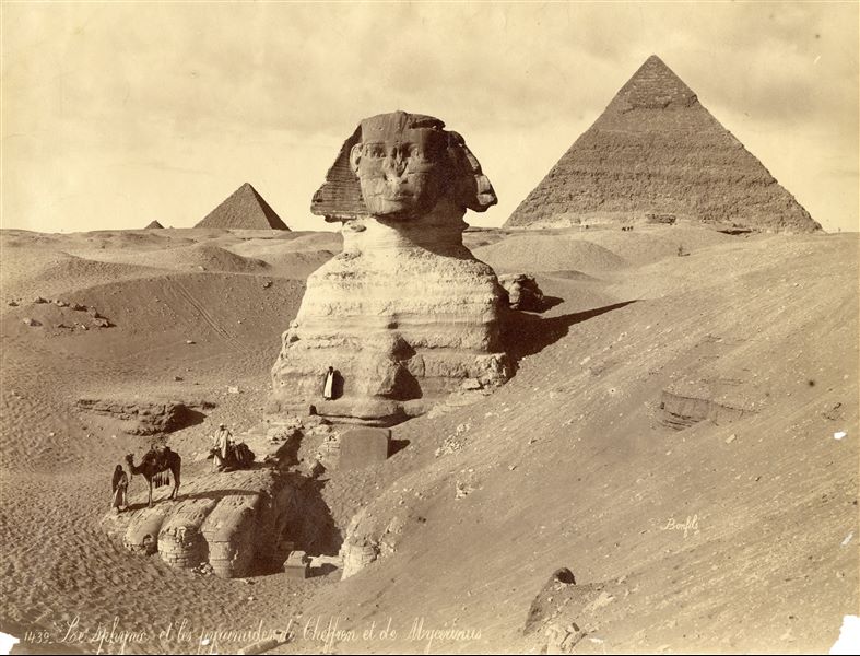 The photograph depicts the Sphinx (still not completely cleared of sand) upon which some egyptians stand. Between its paws, one can clearly see the "Dream Stele", the stele of Pharaoh Tuthmosis IV from the 18th dynasty, as well as some remains of a plinth. In the background, the pyramids of Khafre (right), Menkaure (to the left of the Sphinx's head) and the top of a satellite pyramid in the distance are visible. The author's signature is at the bottom right.  