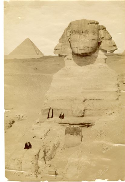 Photograph of the Sphinx in the Giza Plateau, still not completely cleared of sand. The stele of Pharaoh Thutmose IV from the 18th dynasty, covered with mats, and a column base can be seen between the Sphinx's paws. Three men in traditional clothing stand on the monument. In the background on the left, the pyramid of Pharaoh Menkaure can be seen. The signature of the author is at the bottom right.   