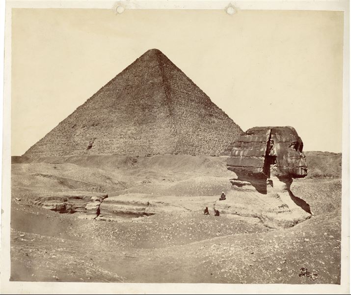 Photograph of the Sphinx in the Giza Plateau viewed from the side (south), still partially buried by desert sands, with three egyptians seated. Behind, the pyramid of Khufu is visible. The image could most likely be attributed to V. R. Lanzone.