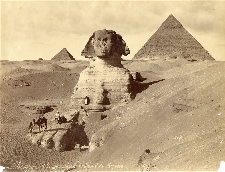 The photograph depicts the Sphinx (still not completely cleared of sand) upon which some egyptians stand. Between its paws, one can clearly see the "Dream Stele", the stele of Pharaoh Thutmose IV from the 18th dynasty, as well as some remains of a plinth. In the background, the pyramids of Khafre (right), Menkaure (to the left of the Sphinx's head) and the top of a satellite pyramid in the distance are visible. The author's signature is at the bottom right.  
