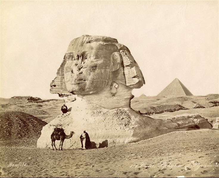 The photograph depicts the Sphinx, partially cleared from sand, with two Egyptians climbing it and another posing with a camel. In the background, two pyramids can be made out, that of Menkaure and a secondary one. The signature of the author can be found at the bottom left.