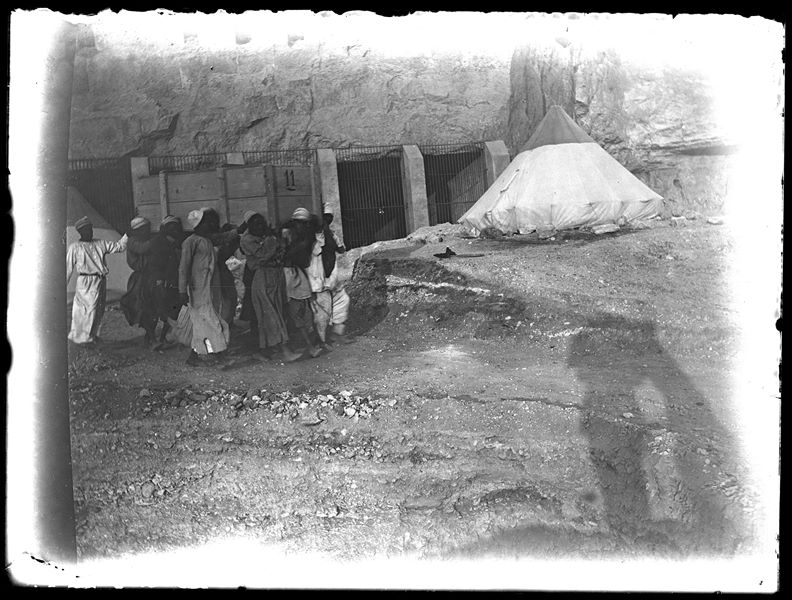  The Italian Archaeological Mission’s camp in front of Tomb IV and Tomb V, which are closed off with gates. In the foreground, a group of Egyptian workmen are carrying a crate, marked with the number 11. The photographer's shadow can be seen at the bottom right. Schiaparelli excavations. 