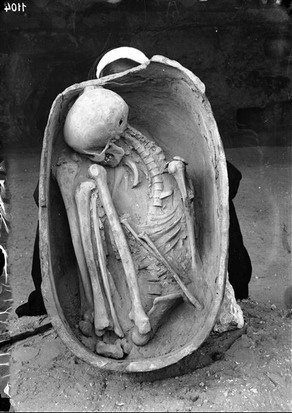 Human remains (secondary burial), placed inside a small terracotta coffin. The coffin is held upright by a worker for it to be photographed. Schiaparelli excavations. 