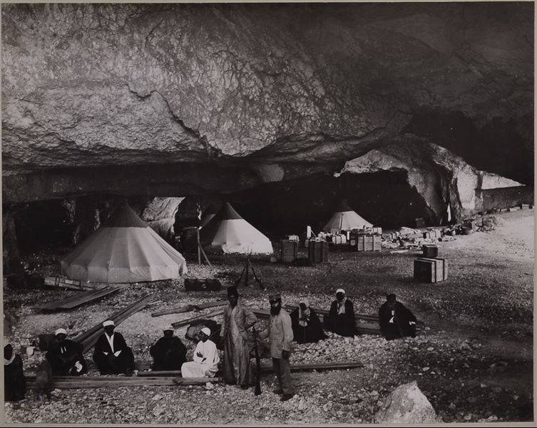 Interior of one of the caves where the Italian Archaeological Mission camped during excavations in the necropoleis of Qau el-Kebir and Hammamiya. In the foreground there are some members of the mission, including the young assistant-cook Buhus dressed in white, the interpreter and guide Bolos Ghattas (in grey), and the mission's “jack of all trades”, Benvenuto Savina, the only one wearing European clothing and holding a rifle. The remaining characters are presumably the cook, Atallah, and the rais (heads of the workers).  In addition to the conical tents, crates for transporting the ancient finds and mission material can be seen in the camp. Photo album, Schiaparelli excavations. 