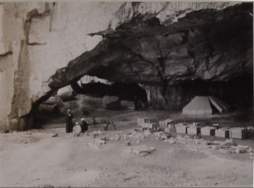 Interior of one of the caves where the Italian Archaeological Mission camped during excavations in the necropoleis of Qau el-Kebir and Hammamiya. The three visible figures are barely recognisable, perhaps the boy dressed in white, due to similarities with other photographs, could be the assistant-cook Buhus, thus suggesting that one of the other two remaining figures is in fact the cook, Atallah. In addition to the conical tents, crates for transporting the ancient finds and mission material can be seen in the camp. Photo album, Schiaparelli excavations. 