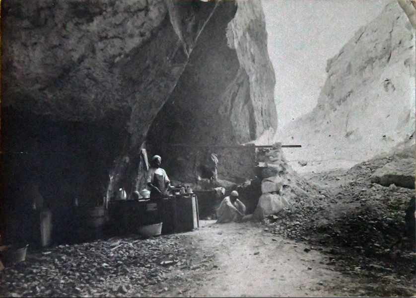 Photograph of the Italian Archaeological Mission’s kitchen in the cave of Qau el-Kebir, where the camp was located. Here, the cook Atallah is preparing some food. Young Buhus, the assistant cook is also present, it appears that he is trying to light the fire. (Original caption: Mission’s kitchen). Angelo Sesana Archive. 