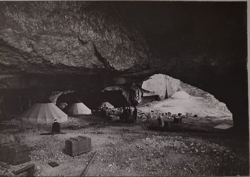 Interior of one of the caves where the Italian Archaeological Mission camped during excavations in the necropoleis of Qau el-Kebir and Hammamiya. The only identifiable individual is the figure in the centre wearing western clothing, undoubtedly Benvenuto Savina. In addition to the conical tents, crates for transporting the ancient finds and mission material can be seen in the camp. Photo album, Schiaparelli excavations. 