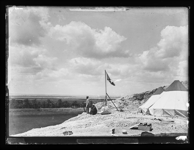  The Italian Archaeological Mission’s camp, with its typical conical-shaped tents, near Qau el-Kebir. Near the Italian flag, a seated Egyptian and Benvenuto Savina look towards the horizon. In the foreground there is a box with archaeological finds. Schiaparelli excavations.