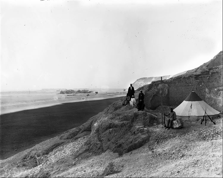 Camp of the Italian Archaeological Mission near the caves of Qau el-Kebir. In the foreground we see Benvenuto Savina and a group of Egyptians; identifiable from the left we see the interpreter and guide Bolos Ghattas (seated), the cook Atallah (standing) with the young assistant Buhus (seated and dressed in light colours). The other two Egyptians are not recognisable. In the background a bridge on a dried-up canal can be seen, presumably the same pictured in B00984. Schiaparelli excavations.