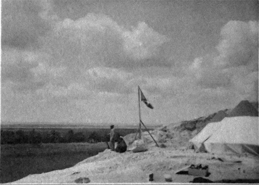 Fotografia di una parte dell’accampamento della Missione Archeologica Italiana durante gli scavi a Qau el-Kebir e Hammamiya nel 1905 e nel 1906. Accanto all’asta con la bandiera italiana, è riconoscibile la figura di Benvenuto Savina, tuttofare della missione. Archivio Angelo Sesana.