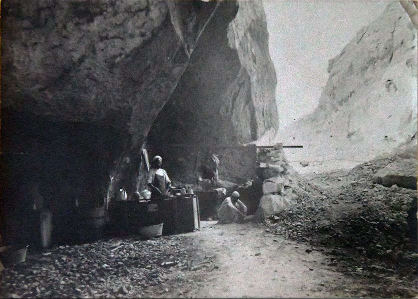 Fotografia della cucina della Missione Archeologica Italiana nella grotta di Qau el-Kebir, dove era situato l’accampamento, in un momento in cui il cuoco, Atallah, sta preparando da mangiare. È presente anche il giovane Buhus, l’aiuto cuoco, forse alle prese con il fuoco. (didascalia originale: Cucina della missione). Archivio Angelo Sesana. 
