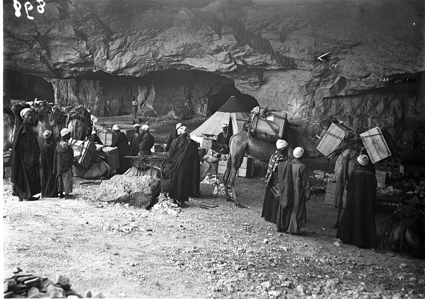 Accampamento della Missione Archeologica Italiana presso le grotte di Qau el-Kebir. In questo scatto, i dromedari sono pronti per partire. Al centro, in via di completamento di una cassa, si riconosce la figura del falegname della missione, Morkos, con un trapano in mano. Sullo sfondo, a sinistra, è visibile Benvenuto Savina. Gli operai egiziani non sono stati riconosciuti. Scavi Schiaparelli.
