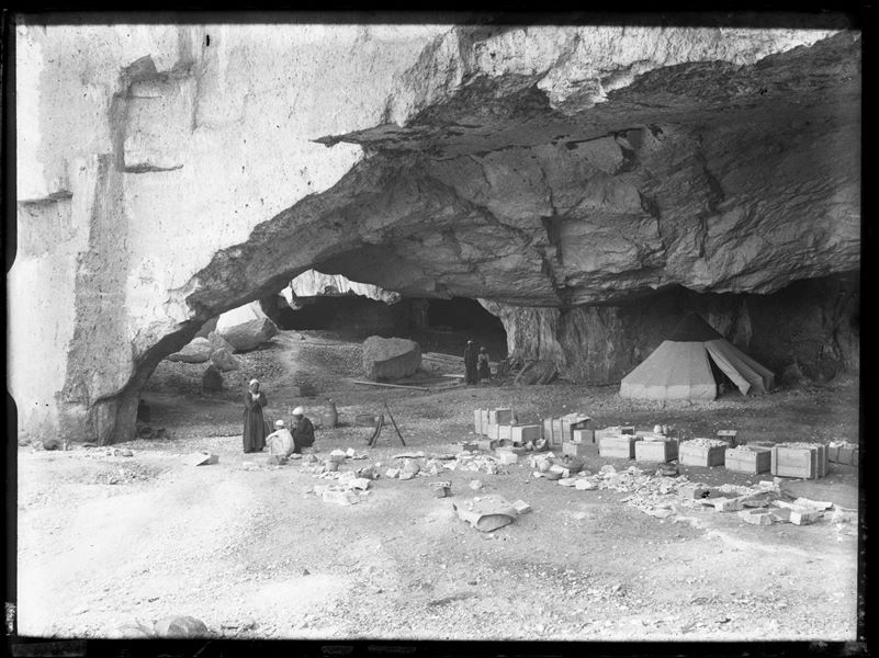 Accampamento della Missione Archeologica Italiana nelle cave di pietra presso Qau el-Kebir. Sono ritratti due gruppi di operai egiziani a riposo o in posa (in fondo, il falegname Morkos e un aiutante). Al fondo vicino alla parete una delle tende della Missione, mentre in primo piano casse contenenti diversi reperti archeologici. Scavi Schiaparelli.