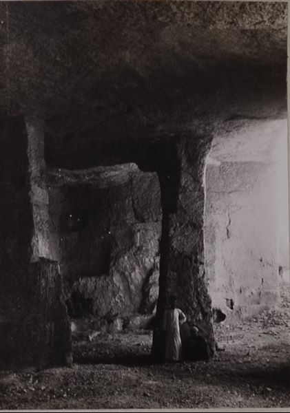 Interior of one of the caves, used as a quarry and then as a camp for the Italian Archaeological Mission, nearby to the rock-cut tombs of Qau el-Kebir. Out of the two figures captured in the image, the one wearing a light-coloured tunic is Bolos Ghattas, the mission's interpretor and guide. Photo album, Schiaparelli excavations.